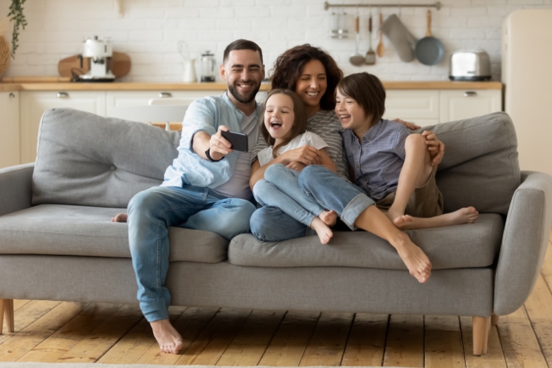 What Are Signs I Need a New Furnace? Smiling parents with little kids laughing using smartphone together sitting on couch at home. Happy father holding phone taking selfie with children. Family watching video having fun with cellphone.